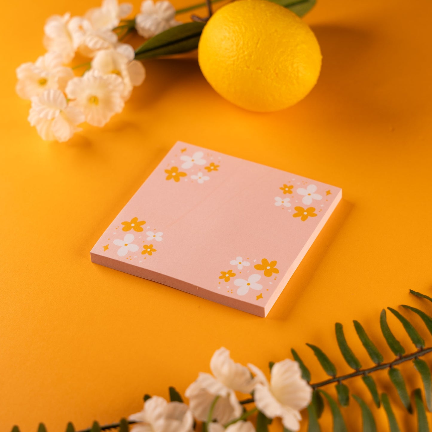 Pink Daisy Sticky Note Pad with daisies in all four corners, displayed on a vibrant yellow background with a lemon, white flowers, and green fern leaves surrounding it